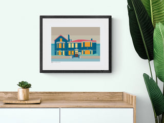 Framed art print of Balham Bowls Club pub above a wooden sideboard with tropical plants.