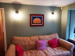 Framed art print of The Bedford Pub in a living room snug. The print hangs on an olive green wall, above a pink sofa with pink cushions.