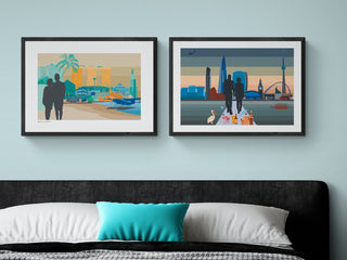 Two bespoke art prints side-by-side on a blue bedroom wall. The prints feature silhouettes of a couple with a skyline of landmarks.