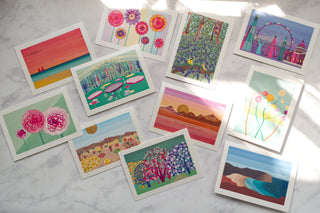 11 illustrated greetings cards scattered on a sunlit table by South Island Art.