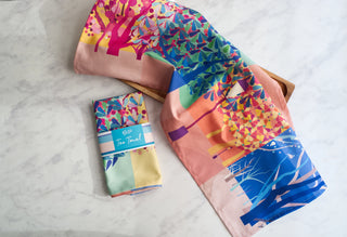 Two folded tea towel with a landscape of trees beside each other. One is in its packaging, the other folded in half, showing the design.