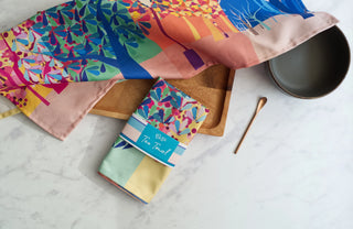 Unfolded tea towel illustrated with trees lies above a folded version with a branded band around it.