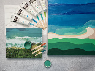 Artist materials including blue and green paint tubes, painted swatch sticks, a painted canvas of an abstract seascape and a photobook with similar colours and ocean scene.