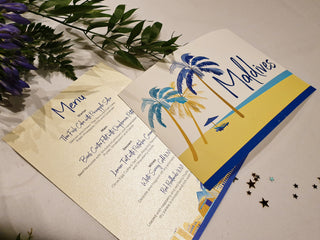 Wedding table name reads Maldives with a seascape illustration. The sign is on top of a printed wedding day menu.