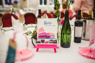 Illustrated table name sign 'Rome' sits on a white table cloth beside bottles of flowers. The sign features an illustration of the colosseum and photos of the couple's holiday.