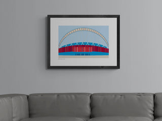 A framed and matted print of Wembley Stadium in red, blue, and white, displayed on a grey sofa.