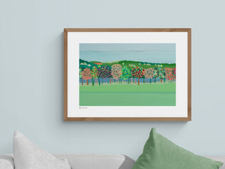A framed and mounted landscape art print featuring a green field with trees in the background, displayed on a wall above a green cushion.