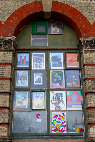 Arched window of Alexandra Palace. Each window pane displays an art print. Three pieces by South Island Art can be seen amongst all the prints.