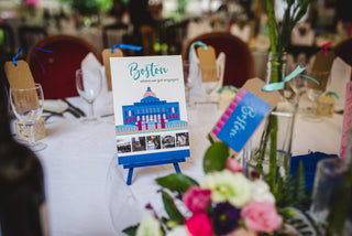 Blue and pink wedding table name in situ, surrounded by flowers and glasses. The table name reads Boston and has an illustration of the Massachusetts State House with photos of the bride and groom.