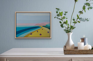 Painted Mediterranean coastal landscape in a natural wooden floater frame above a wooden dining table.