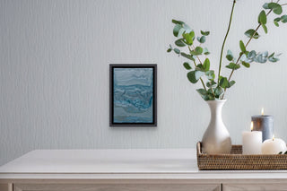 Framed grey and turquoise fluid painting on a white textured wall above a dining table with vase and candles.