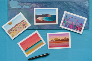 5 greetings cards, each illustrated with a different landscape, sit on top of a painted board and turquoise background.