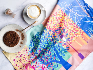 Colourful tea towel illustrated with trees laid out on table with a plate of cookies and a cappuccino