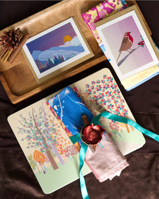 A selection of gifts including placemat illustrated with autumnal trees, rolled tea towel in blue and pink tied with a turquoise ribbon and Christmas cards designed with a robin and winter forest