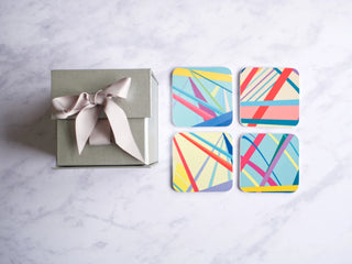 Silver gift box with ribbon beside set of four multicoloured coasters each with a different abstract design.