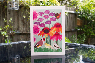 Illustrated greetings card of summer birds, butterflies and flowers. The card is standing on an outdoor table in a garden.